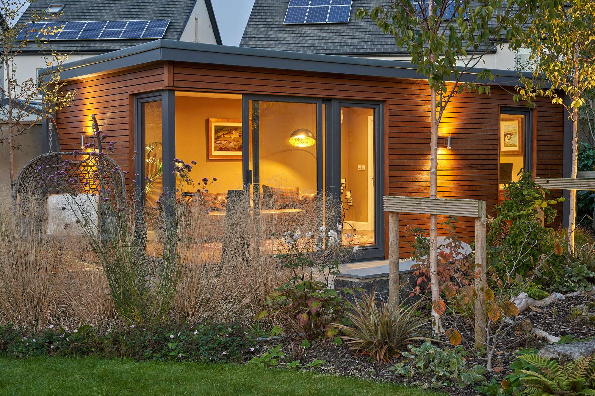 A cedar-clad Shomera Classic Range garden studio lit at dusk, its corner sliding doors glowing over a planted garden