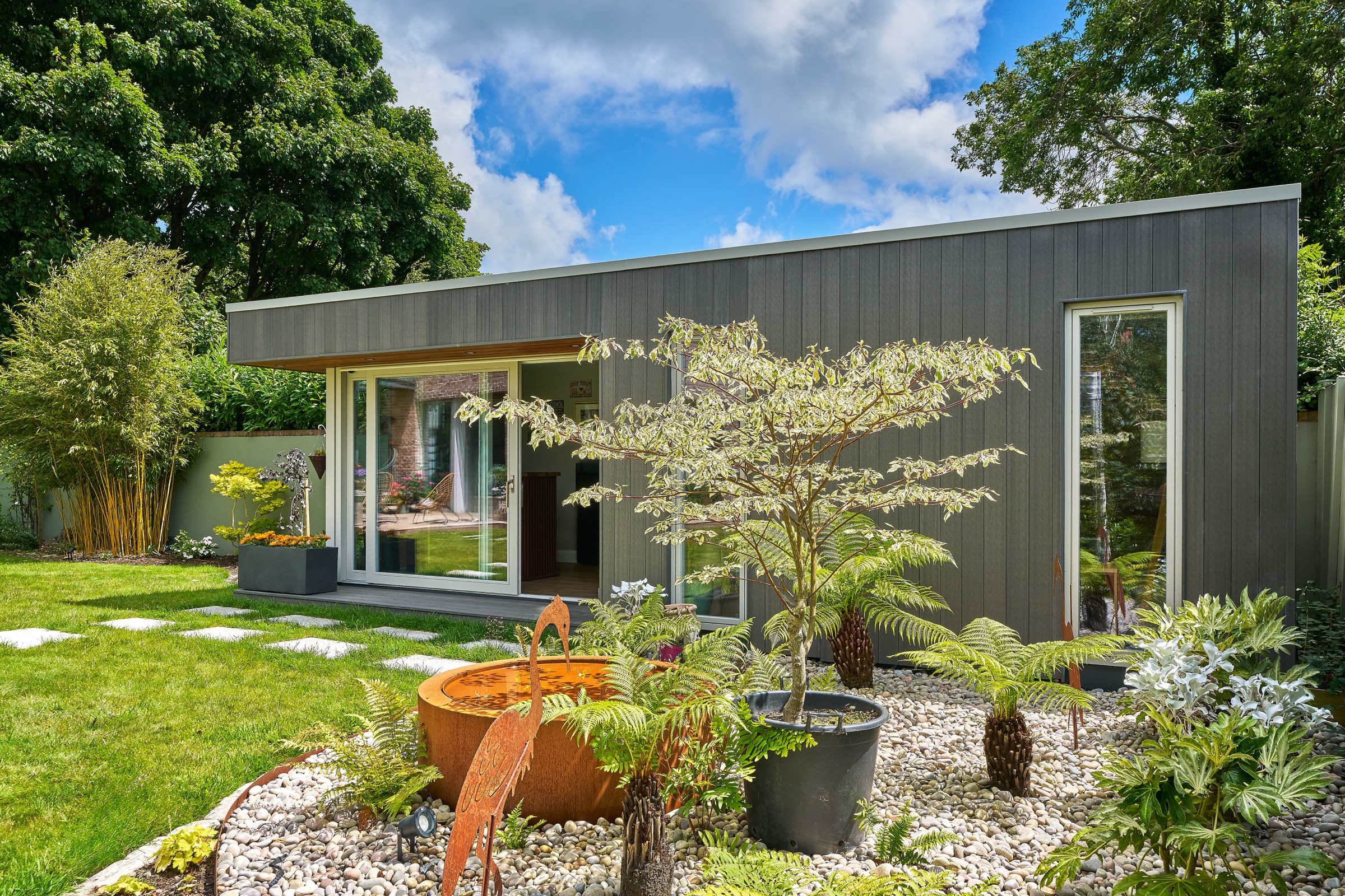A grey timber-clad Shomera garden studio in a richly planted garden on a sunny day