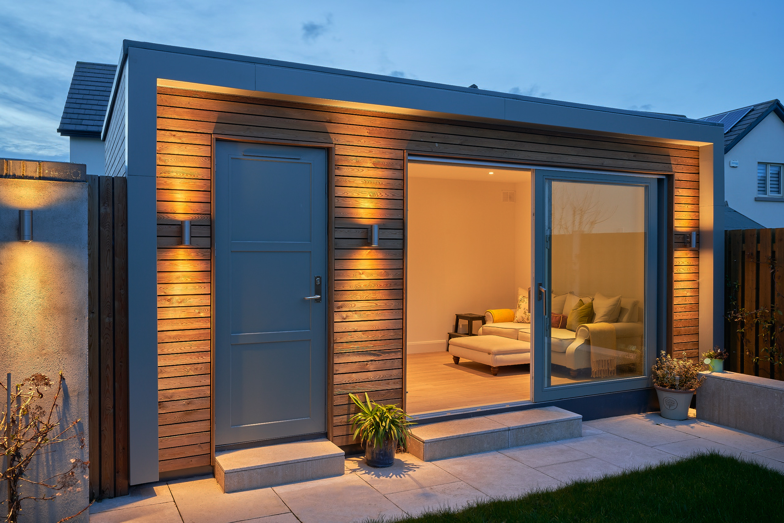 The cedar-clad garden studio at dusk, uplighters washing the cladding and the lounge lit through open sliding doors