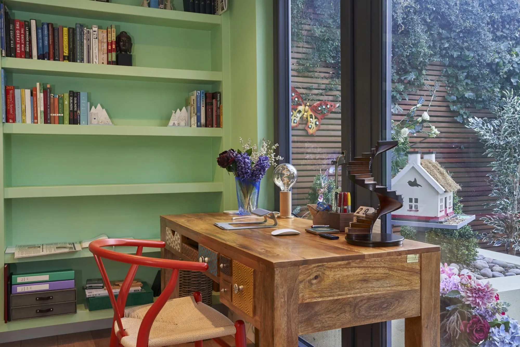 A desk and a red chair in a garden room with green built-in bookshelves and floor-to-ceiling windows