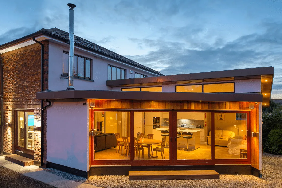 A house extension with a flat overhanging roof and a wall of timber-framed glazing, lit at dusk