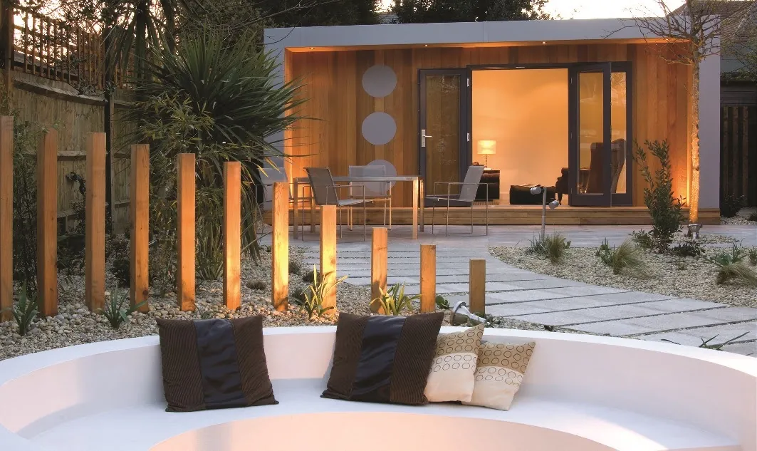 A cedar-clad garden room at dusk, seen across a gravel garden with timber posts and a curved white seat