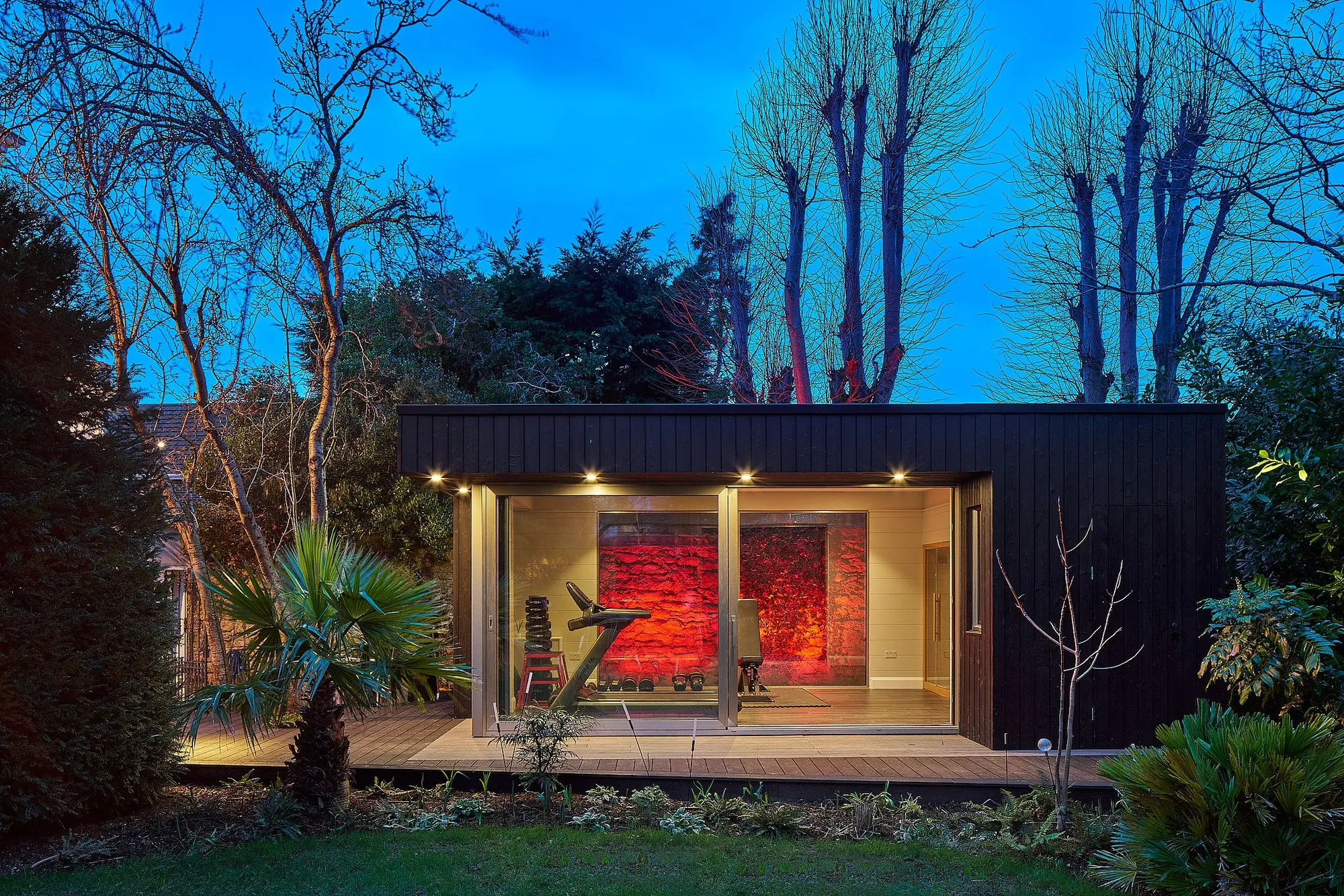 A black-clad garden room used as a home gym, lit at dusk among trees