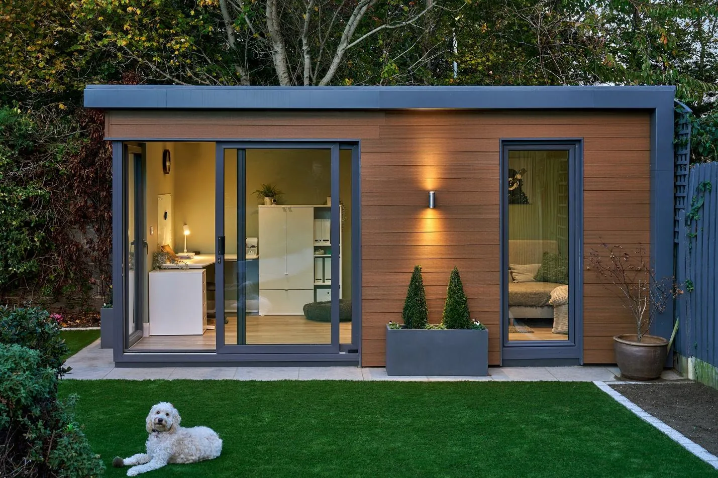 A timber-clad garden room with sliding glass doors on a lawn, with a white dog lying on the grass