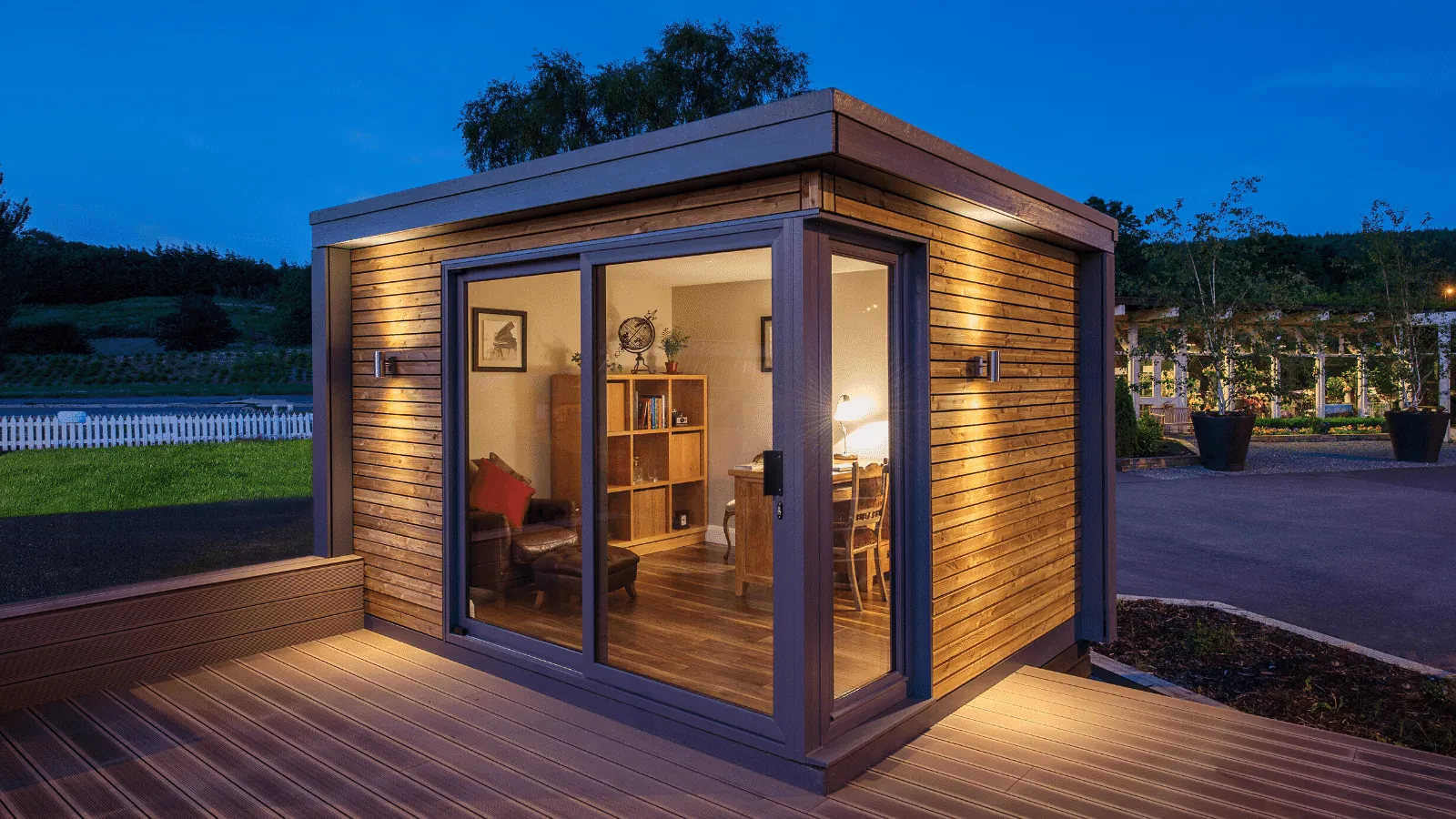 A small timber-clad garden room with sliding glass doors on a deck, lit at dusk