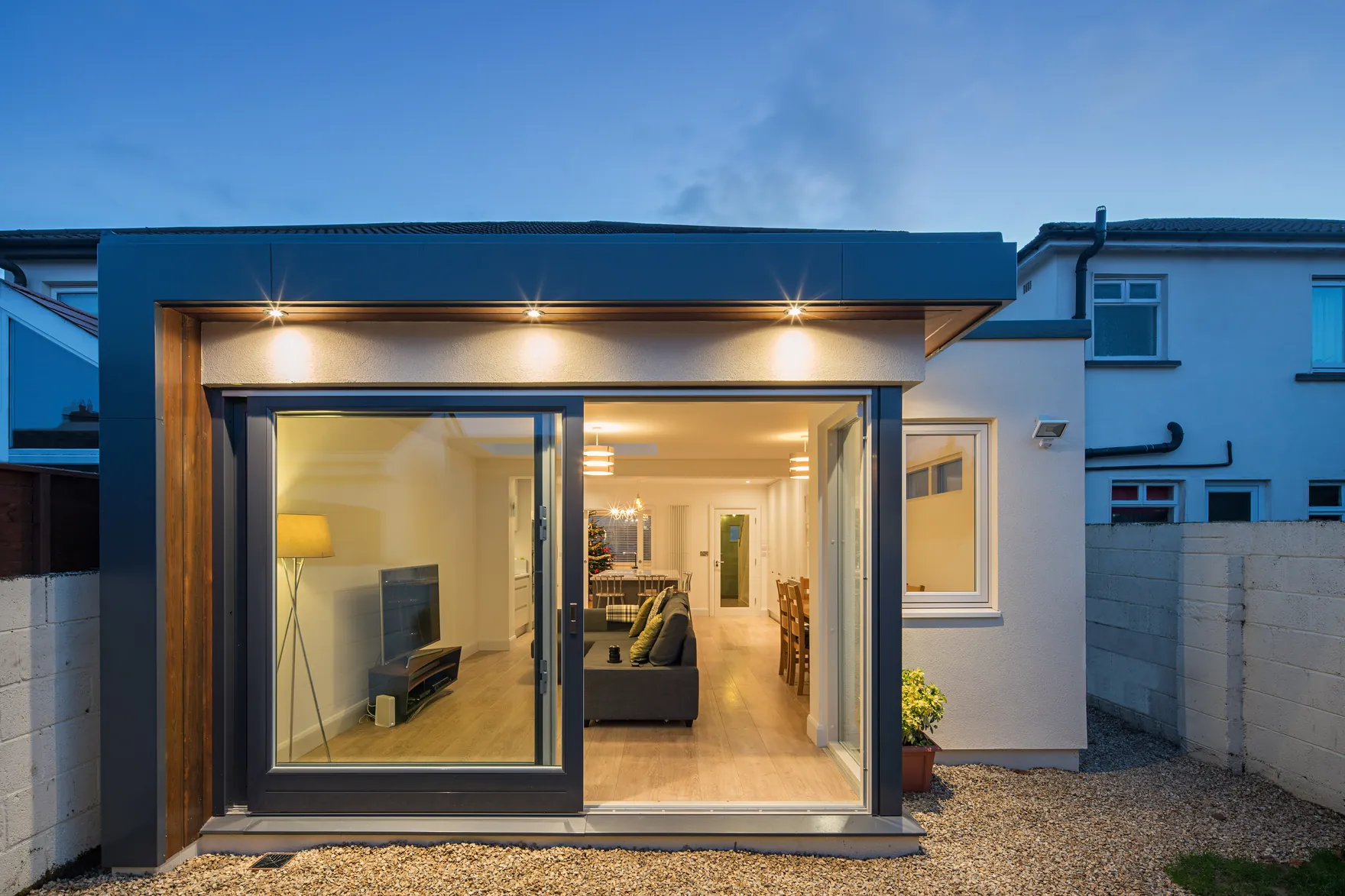 A dark-clad garden room with sliding glass doors open onto a living room, lit at dusk