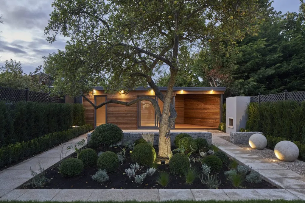 A cedar-clad garden room with a covered seating area behind a mature tree and clipped planting, lit at dusk